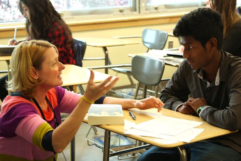 A female teacher kneeling in front of student's desk explaining.