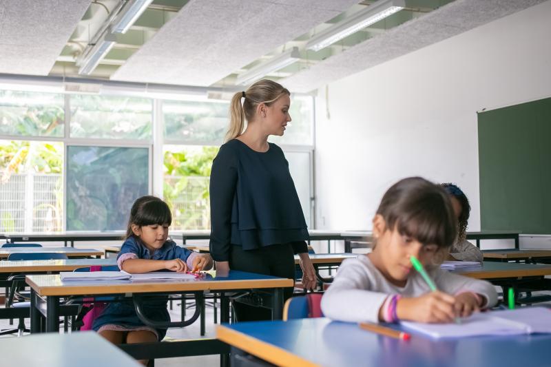 A female teacher standing over and observing students working on their tasks.